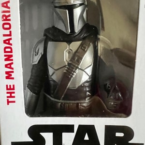 Puede incluir: Figura de acci&oacute;n de The Mandalorian en un embalaje de pl&aacute;stico transparente. La figura es plateada y negra, con casco y armadura. El logotipo de Star Wars es visible en la parte inferior del embalaje, y "THE MANDALORIAN" est&aacute; impreso en el lateral.