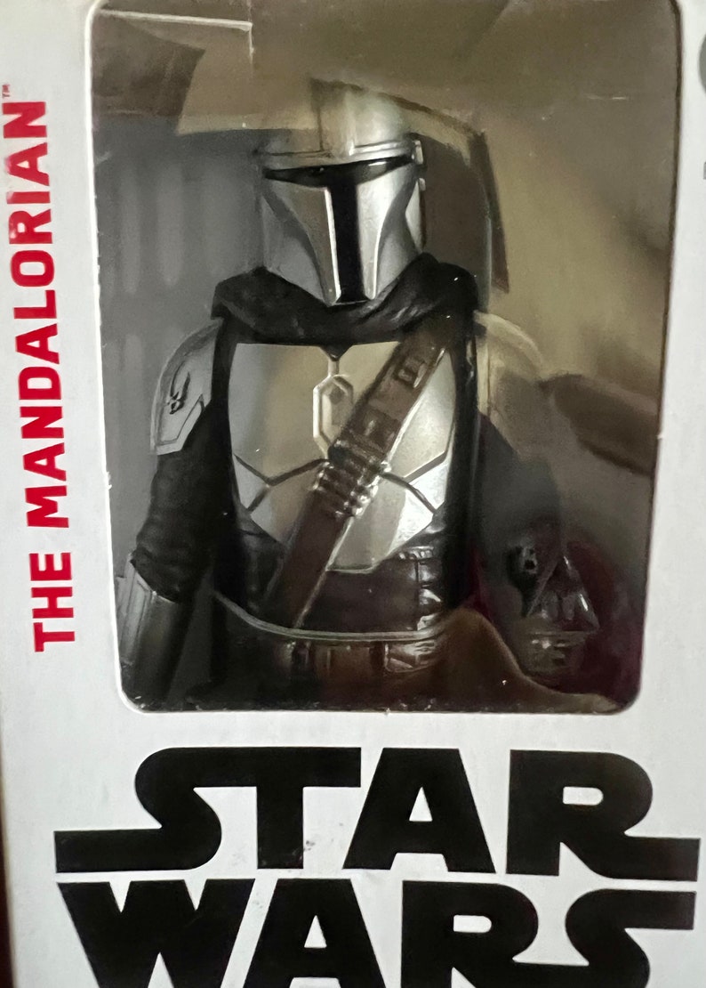 Puede incluir: Figura de acci&oacute;n de The Mandalorian en un embalaje de pl&aacute;stico transparente. La figura es plateada y negra, con casco y armadura. El logotipo de Star Wars es visible en la parte inferior del embalaje, y "THE MANDALORIAN" est&aacute; impreso en el lateral.