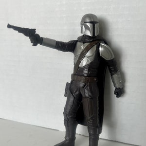 Puede incluir: Una figura de acci&oacute;n detallada de The Mandalorian, un personaje de la franquicia Star Wars. La figura est&aacute; vestida con una armadura plateada y negra, sosteniendo una pistola bl&aacute;ster. La figura est&aacute; posada como si apuntara el arma.
