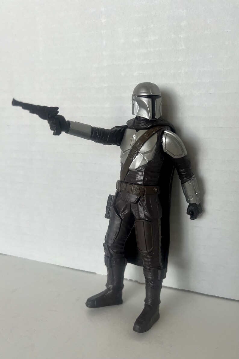 Puede incluir: Una figura de acci&oacute;n detallada de The Mandalorian, un personaje de la franquicia Star Wars. La figura est&aacute; vestida con una armadura plateada y negra, sosteniendo una pistola bl&aacute;ster. La figura est&aacute; posada como si apuntara el arma.