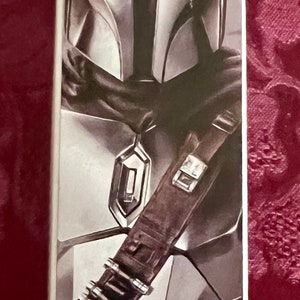 Puede incluir: Caja vertical con una ilustraci&oacute;n de The Mandalorian de Star Wars. La imagen muestra el casco y la armadura del personaje en plata y gris. El logotipo de Disney est&aacute; en la parte inferior de la caja.