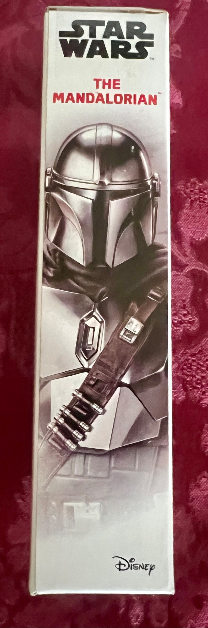 Puede incluir: Caja vertical con una ilustraci&oacute;n de The Mandalorian de Star Wars. La imagen muestra el casco y la armadura del personaje en plata y gris. El logotipo de Disney est&aacute; en la parte inferior de la caja.