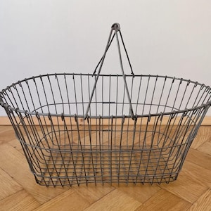 Puede incluir: Una cesta de alambre de estilo vintage con base rectangular y parte superior redondeada. La cesta está hecha de alambre de metal gris y tiene un asa que se arquea sobre la parte superior. La cesta está sobre un suelo de madera.