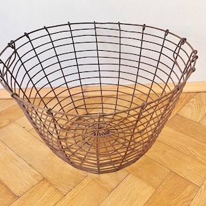 Puede incluir: Una cesta grande de alambre de tejido abierto con forma redondeada. La cesta es de metal marrón oscuro y tiene un patrón de rejilla. La cesta está vacía y descansa sobre un suelo de madera.