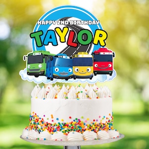 Puede incluir: Un adorno para pastel de cumpleaños con cuatro autobuses de dibujos animados con caras sonrientes. El adorno está decorado con un círculo azul con el texto "Happy 2nd Birthday Taylor".