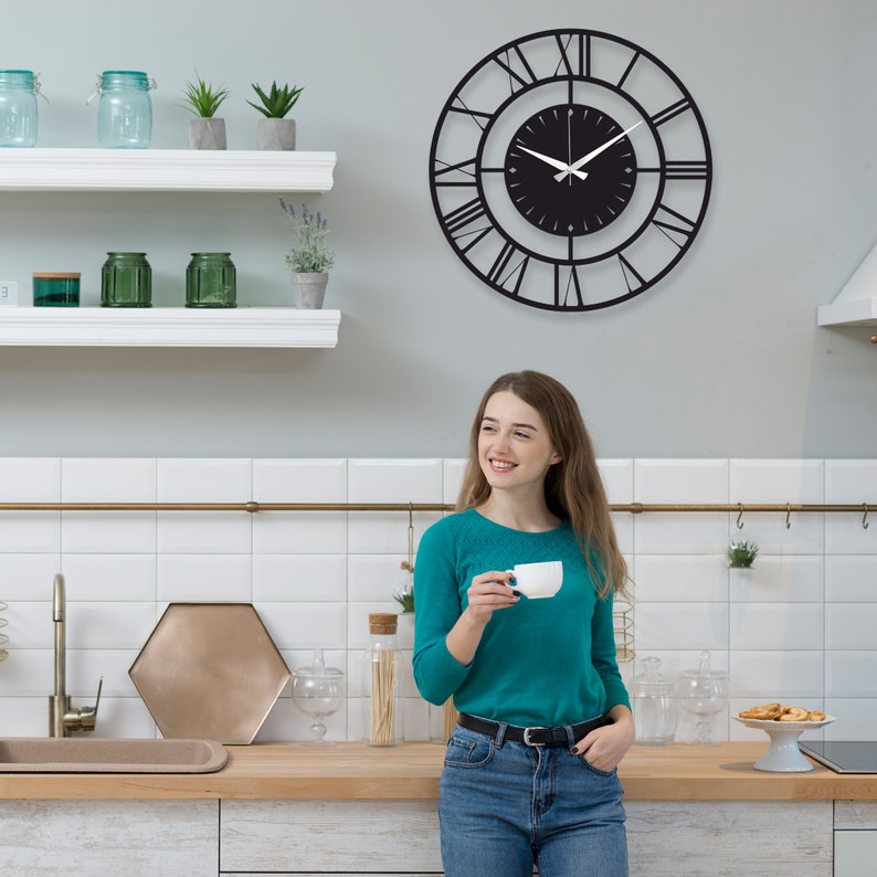 Może przedstawiać: Duży, czarny, okrągły zegar ścienny z rzymskimi cyframi wisi na jasnoszarej ścianie. Zegar ma minimalistyczny design z czarną tarczą i białymi wskaz&oacute;wkami. Zegar jest centralnym punktem w kuchni.