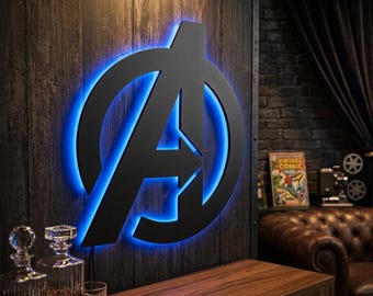 May include: A matte black metal wall sign featuring the Avengers logo, illuminated from behind with a vibrant blue LED light. The circular emblem with the stylised letter A is mounted on a dark wood panel, creating a glowing blue silhouette.