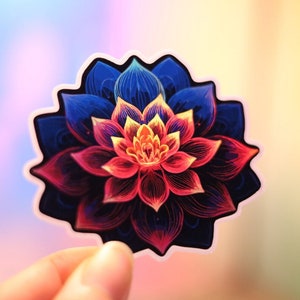 May include: A colorful sticker featuring a stylized lotus flower. The flower has layered petals in shades of blue, red, and yellow, with a dark outline. The sticker is held against a blurred, colorful background.