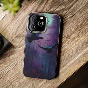 May include: A smartphone case featuring a dark, artistic design of crows in flight over a forest. The case has a purple, blue, and teal color scheme, with a glossy finish. The case is shown on a wooden surface.