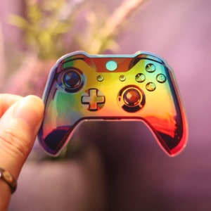 May include: A holographic sticker in the shape of a video game controller. The sticker has a rainbow color scheme and features buttons, a directional pad, and control sticks. The sticker is held up by a hand.