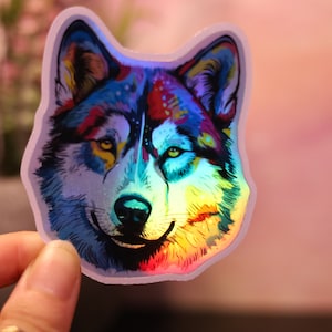May include: A holographic sticker of a wolf's head, with a colourful design. The wolf's fur is rendered in blues, reds, yellows, and greens, giving an iridescent appearance. The sticker has a white border and a smiling expression.