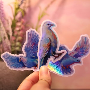 以下が含まれることがあります： 水彩画で描かれたハトのイラストが描かれた3つのステッカー。ハトは主に青色で、虹色の羽とオレンジと紫のアクセントがあります。ステッカーは白い縁取りがあり、柔らかなピンク色の背景に配置されています。