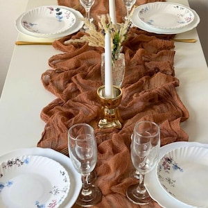 May include: A brown gauze table runner with a textured surface. The runner is laid out on a white table with white plates, gold silverware, and clear glasses. The text "BROWN" is printed on the runner.