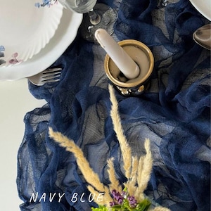 Può includere: Un runner da tavolo blu navy con un tessuto strutturato e stropicciato. Il runner è drappeggiato su un tavolo e ha un mortaio e un pestello decorativi al centro.