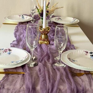 May include: A purple gauze table runner with a gold metal candle holder and two white candles. The runner is laid out on a white table with two white plates with floral designs and two gold forks. The text "LAVANDER" is printed on the runner.