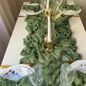 May include: A green table runner with a ruffled texture, placed on a white table. Two white plates with floral designs are set with green napkins, gold cutlery, and clear wine glasses. A white candle in a gold holder is centred on the runner.