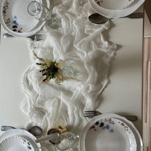 May include: A white table runner with a delicate, textured weave, laid over a white table. Two sets of white plates with floral designs are set on the table, along with glasses and silverware.