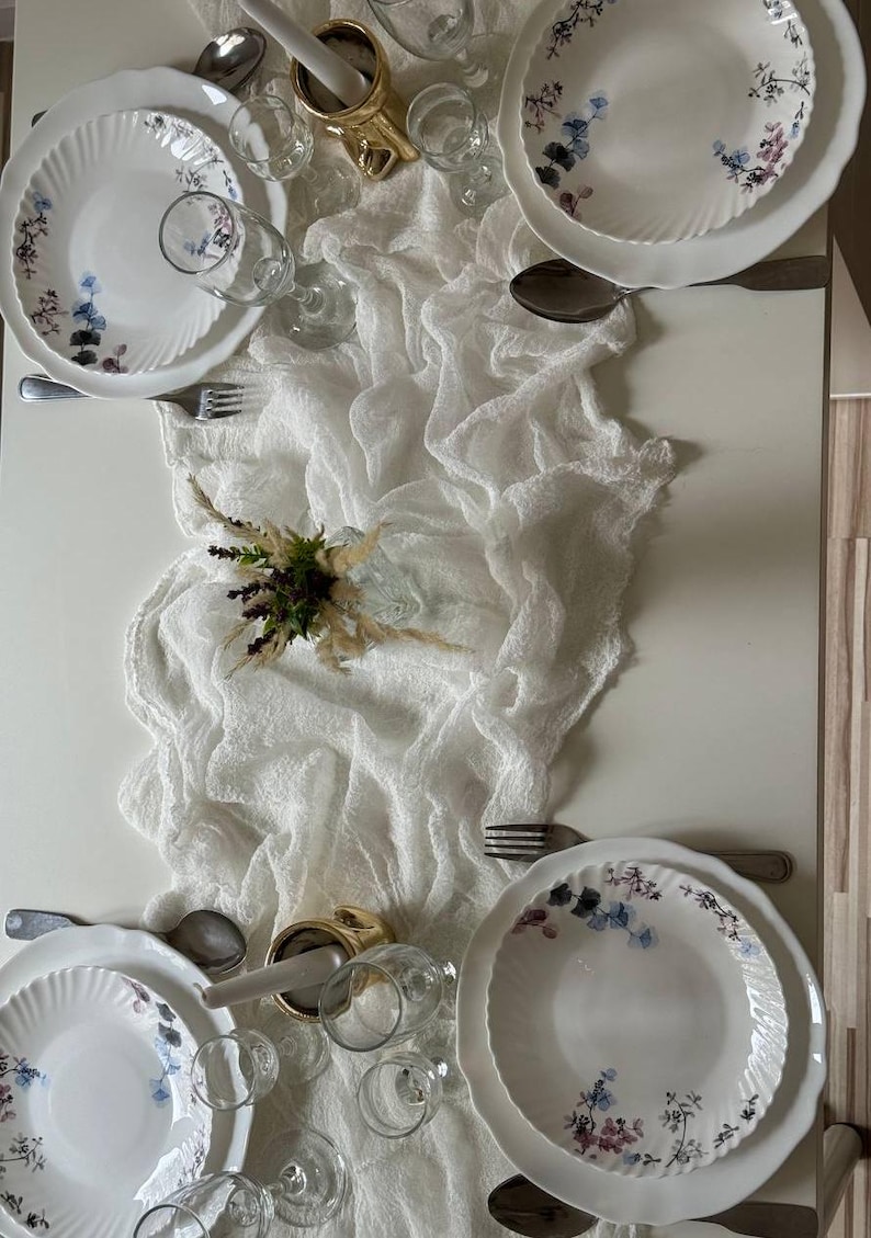 May include: A white table runner with a delicate, textured weave, laid over a white table. Two sets of white plates with floral designs are set on the table, along with glasses and silverware.