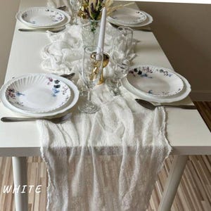 May include: White table runner with a delicate, gauzy texture. The runner is laid over a white table and features a floral pattern on the plates.