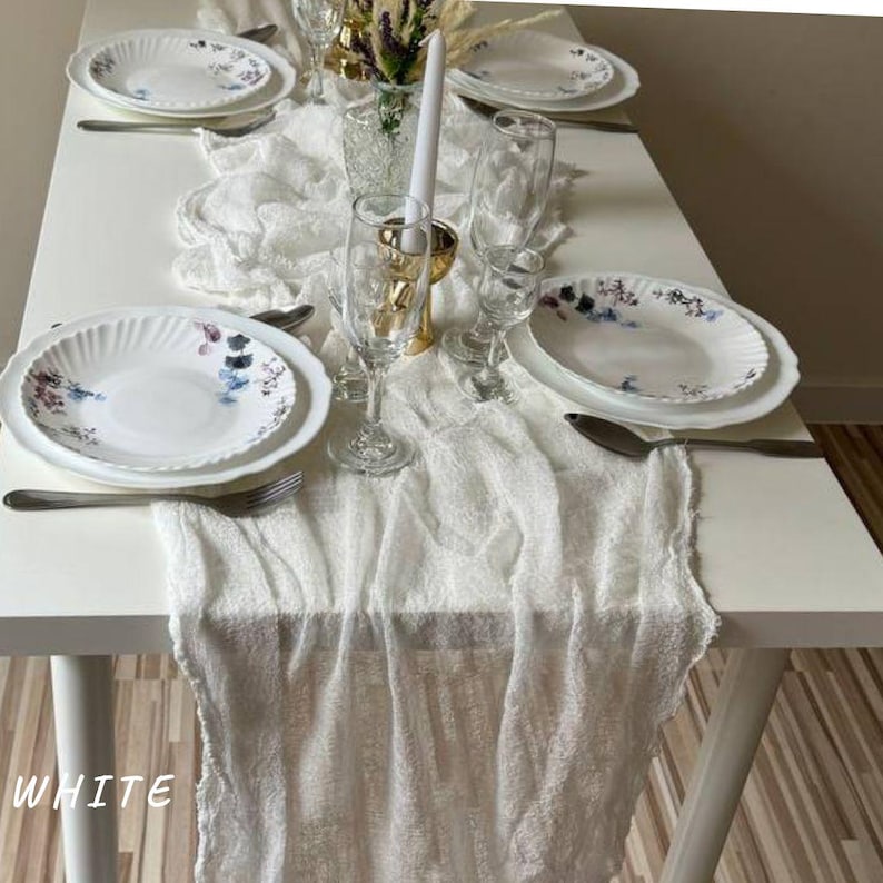 May include: White table runner with a delicate, gauzy texture. The runner is laid over a white table and features a floral pattern on the plates.