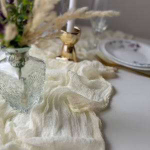 May include: A white sheer table runner with a delicate texture, ideal for adding a touch of elegance to any table setting. The runner is draped over a table with a vase of purple flowers and a gold candle holder.