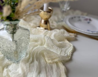 May include: A white sheer table runner with a delicate texture, perfect for adding a touch of elegance to any table setting. The runner is draped over a table with a vase of purple flowers and a gold candle holder.