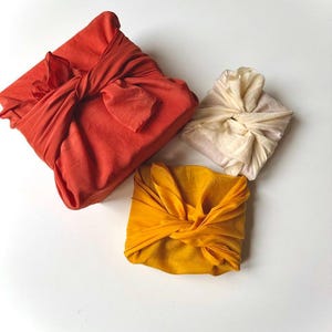 Puede incluir: Tres regalos envueltos en tela de color naranja, amarillo y blanco. Los regalos están atados con tela y están dispuestos sobre una superficie blanca.