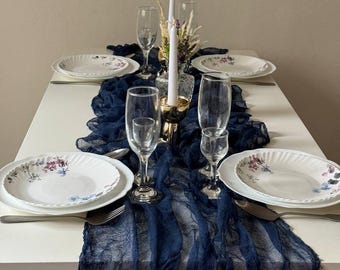 May include: A navy blue table runner with a distressed look, placed on a white table with white plates, gold candle holders, and clear glasses. The runner is made of a soft, sheer fabric and has a slightly ruffled edge.