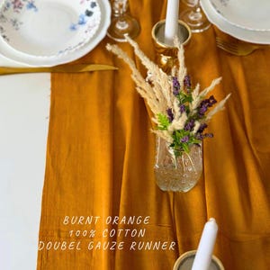 May include: Burnt orange double gauze table runner made of 100% cotton. The runner is displayed on a white table with a vase of dried flowers and candles.