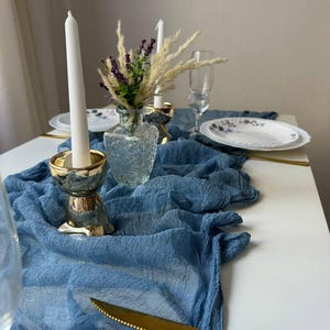 Op de afbeelding: Een blauwe gaas tafelrunner met twee gouden kaarsenstandaards en een heldere glazen vaas met gedroogde bloemen en witte kaarsen.