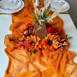 Può includere: Un runner da tavolo arancione con un centrotavola di girasoli, lavanda secca e piccole zucche. Il runner è realizzato in un tessuto trasparente e vaporoso.