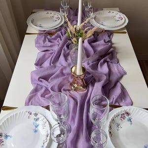 May include: A purple table runner with a white candle and floral centerpiece. Two white plates with floral designs are on either side of the centerpiece. There are four clear glasses on the table.