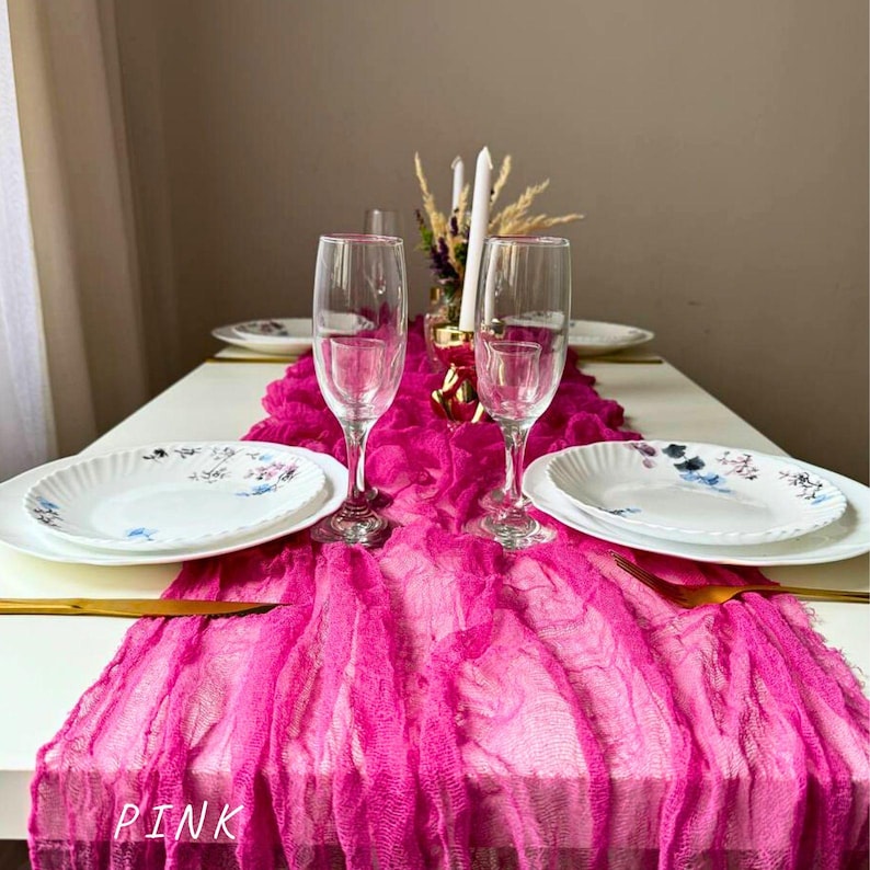 May include: A pink table runner with a ruffled texture, placed on a white table with two clear wine glasses and two white plates with floral designs. The table runner is labeled "PINK".