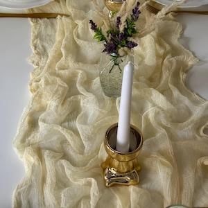 May include: A table setting featuring a cream-colored, textured table runner. Gold candle holders with white candles are placed on the runner, along with a small glass vase with purple flowers and decorative grasses. White plates with floral designs are also visible.