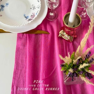 Puede incluir: Camino de mesa de doble gasa rosa hecho de 100% algodón. El camino de mesa es rosa y tiene un sutil efecto degradado. Se muestra en una mesa con platos blancos, cubiertos dorados y un candelero dorado.