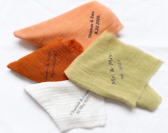 May include: Four folded fabric napkins with frayed edges in peach, burnt orange, olive green, and white. Each napkin features black printed text: Thomas & Ewa 8.22 2026, Emma & Noah 05 June 2027, Mr. & Mrs. est 2027, and Charlote & James 22 May 2027.