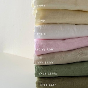 May include: A stack of folded fabric in various colors, including ivory, sand, white, pastel pink, light beige, sage green, and sage gray. The names of the colors are printed on the fabric.