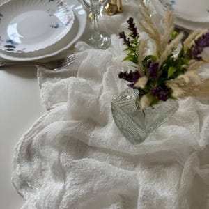 May include: White table runner with a delicate, textured weave. A white plate with blue floral designs sits on the runner, along with a glass of water and a vase of purple flowers.