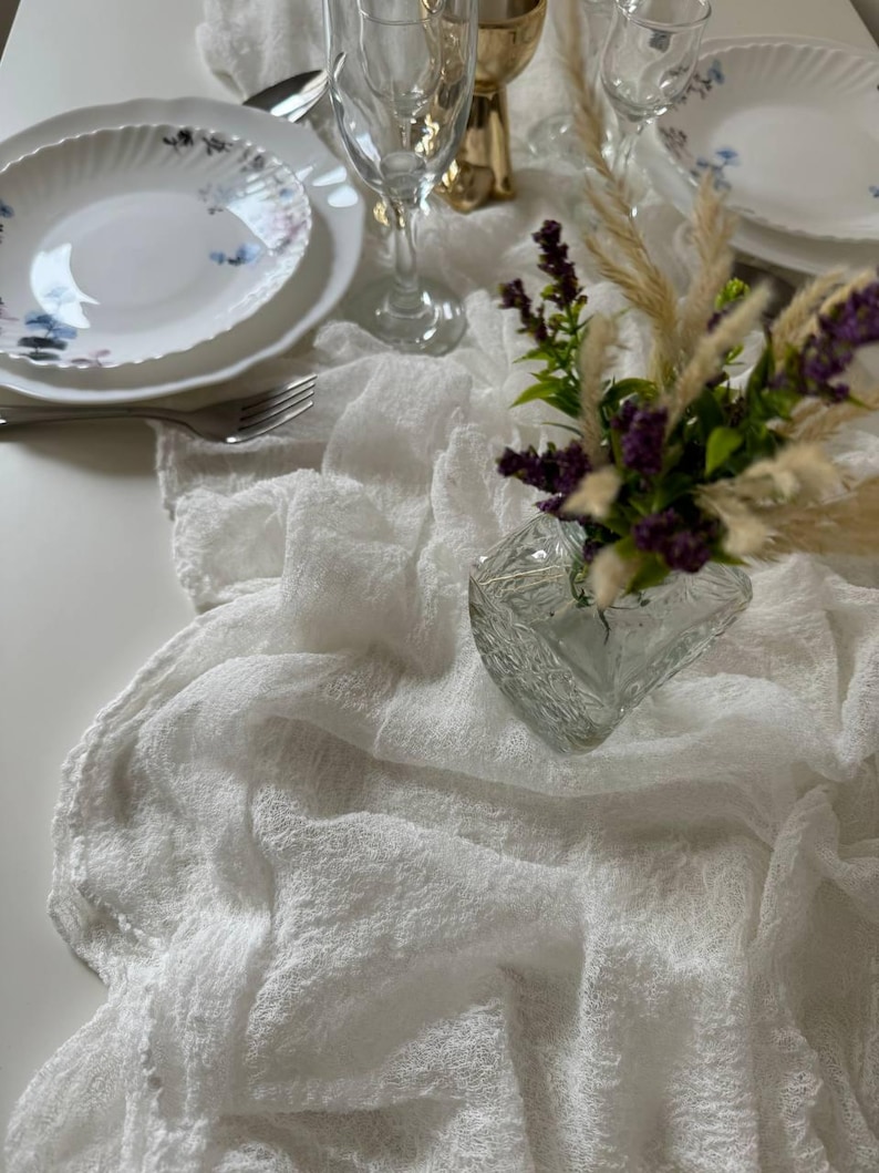 May include: White table runner with a delicate, textured weave. A white plate with blue floral designs sits on the runner, along with a glass of water and a vase of purple flowers.