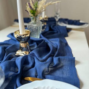 Könnte beinhalten: Ein blauer Tischläufer aus Leinen mit einem goldenen Kerzenhalter und einer weißen Kerze. Der Läufer liegt auf einem weißen Tisch und im Hintergrund steht eine Vase mit Trockenblumen.