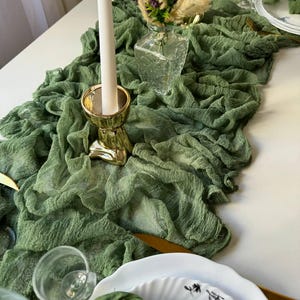 May include: A green gauze table runner with a white candle in a gold candlestick, a clear glass vase with purple flowers and dried grasses, and a white plate with a black floral design.