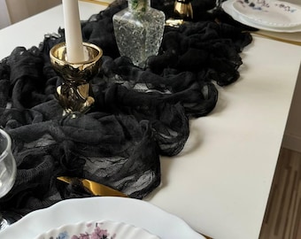 May include: Black sheer table runner with gold candle holders and a clear glass vase with dried flowers. The table runner is on a white table with white plates and gold silverware.