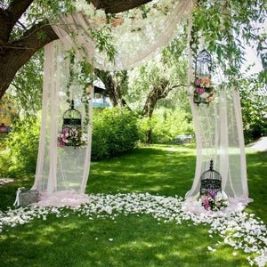Peut inclure: Une arche de mariage extérieure décorée de tissu rose transparent, de fleurs et de cages à oiseaux suspendues remplies de fleurs. La scène se déroule sur une pelouse verte parsemée de pétales de fleurs blanches, sous un grand arbre.