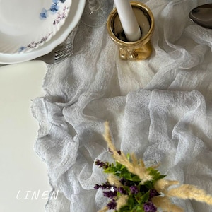 May include: A light gray linen table runner with a delicate texture. The runner is draped over a white table and has a subtle sheen.