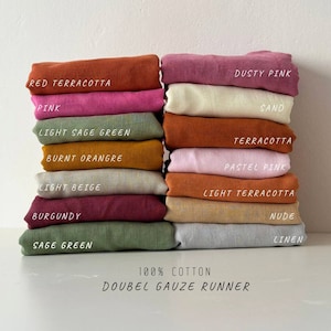 Op de afbeelding: Een stapel van 100% katoen dubbelgaas stofstalen in verschillende kleuren, waaronder rood terracotta, roze, licht saliegroen, gebrand oranje, licht beige, bordeauxrood, saliegroen, stoffig roze, zand, terracotta, pastelroze, licht terracotta, nude en linnen. De tekst "100% KATOEN DUBBELGAAS LOPER" is onder de stofstalen gedrukt.