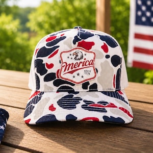 May include: A baseball cap featuring a camouflage pattern in red, white, and blue. The front panel displays a shield-shaped patch with an eagle graphic, the text 'Merica', and 'SINCE 1776' with three stars below the text, all centered on the crown.