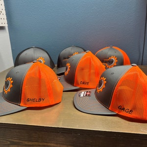 May include: Five grey and orange baseball caps with mesh backs. The caps have a black gear logo with the text "Auto Repair" and a name embroidered on each cap: Shelby, Dave, Gage, and two others.