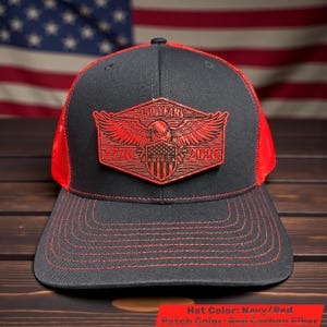 May include: Navy and red trucker hat with a red patch featuring an eagle, shield, and the text "250 YEARS 1776-2026 MADE IN AMERICA". The hat has a mesh back and a curved brim with red stitching.