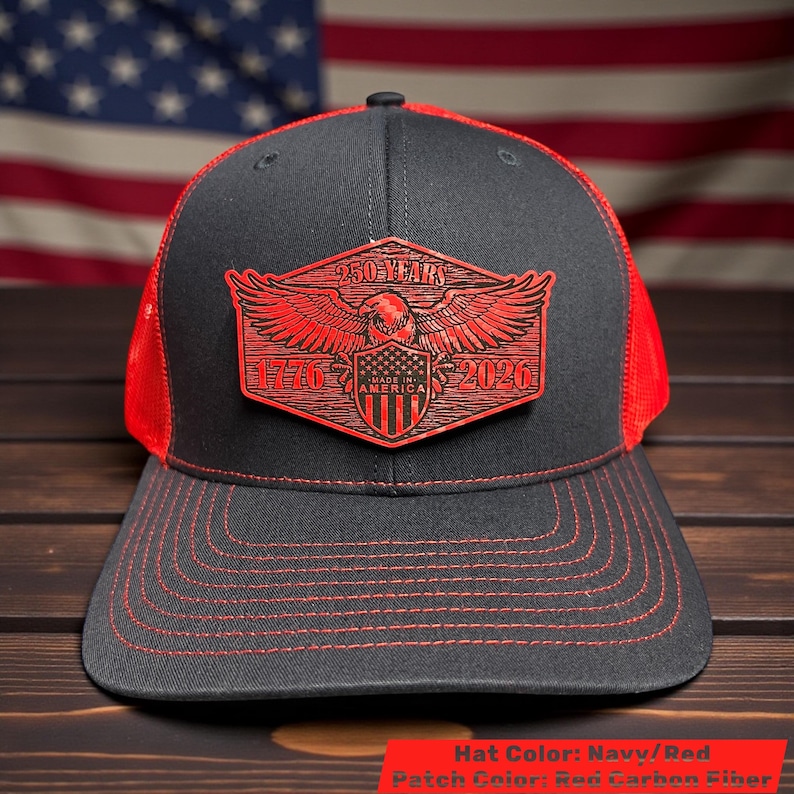May include: Navy and red trucker hat with a red patch featuring an eagle, shield, and the text "250 YEARS 1776-2026 MADE IN AMERICA". The hat has a mesh back and a curved brim with red stitching.