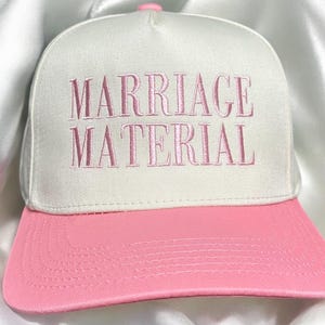 May include: A white baseball cap with a pink brim. The cap has the words "Marriage Material" embroidered in pink on the front.